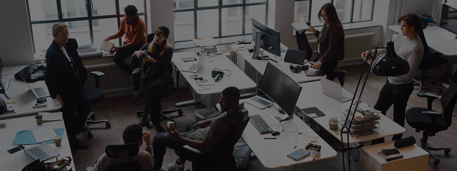 Young creative men and women office workers at their messy desks talking to each other in brightly lit office with three large oversized windows behind them