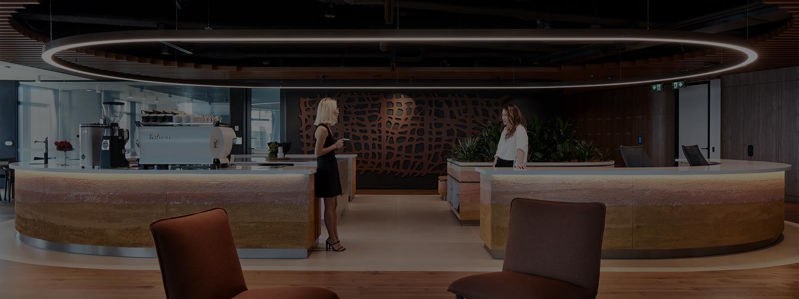 Two young women are standing facing each other on a low-lit hotel-style office floor in front of a long, rounded barista bar. There is a large oval light in the wood-panelled ceiling above their heads. In the foreground are two soft empty chairs and a small coffee table on a round rug, on a wooden floor.