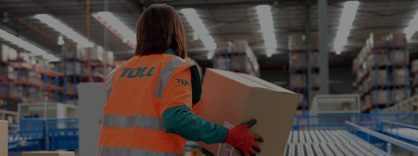 Woman in high vis jacket with ‘TOLL’ written on the back. Her back is facing the camera and she is in a warehouse moving boxes