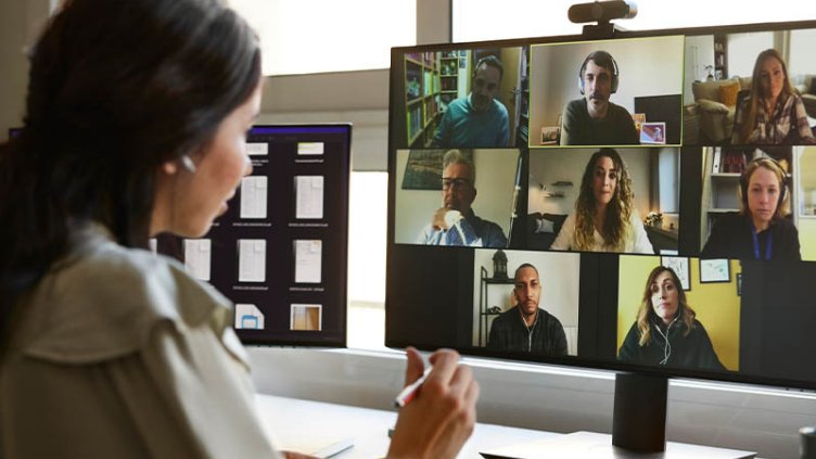 Woman with long brown hair working at her home desk, on a video call with colleagues on her screen