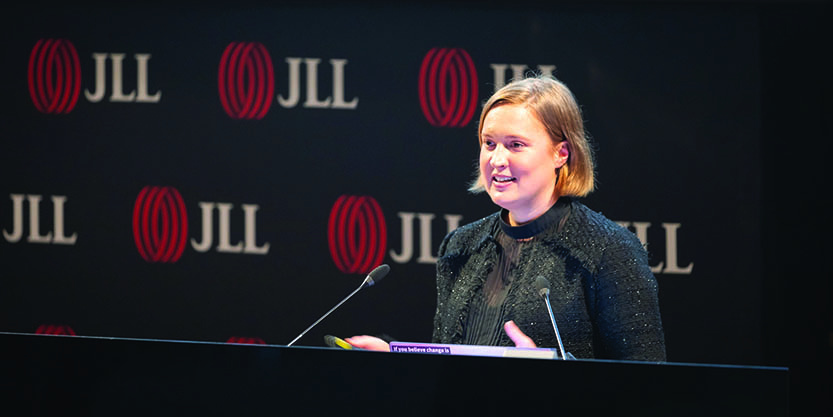 Caitlin Uren, a lady with blonde bobbed hair, wearing a black top, standing at a lectern with two microphones in front of her, presenting. The JLL logo appears in the backdrop