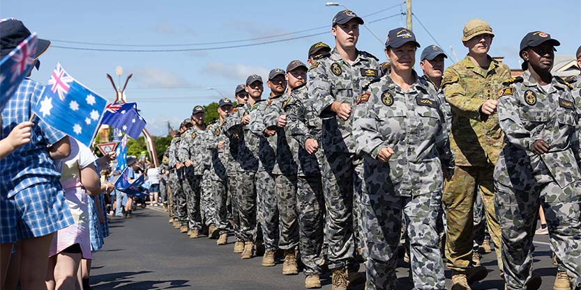 Rows of uniformed Defence personnel march on a road, watched by schoolchildren waving Australian flags
