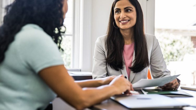 Two professional women engage in a conversation while sitting together in a modern open office space