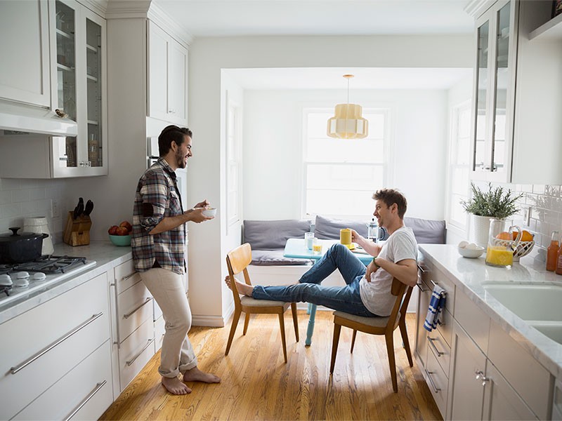 2 male talking to each other at home(one sitting on chair with cup of coffee and other is standing with a bowl of food)