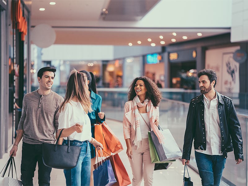 A group of boys and girls shopping together inside a bustling shopping center. They're browsing stores, carrying bags, and chatting