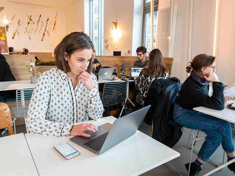 A female sitting on a chair and working on her laptop putting on table (other employees sitting beside and behind her and working)