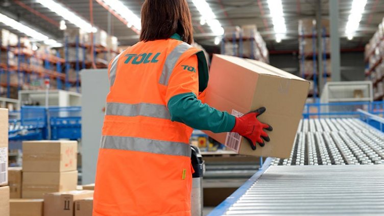 Woman in high vis jacket with ‘TOLL’ written on the back. Her back is facing the camera and she is in a warehouse moving boxes